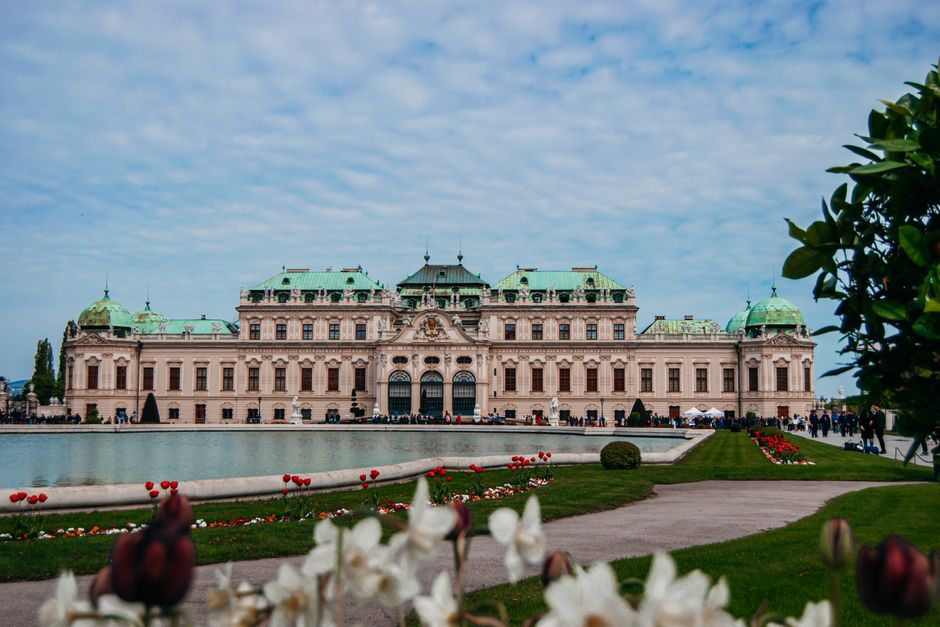 A grand historic building with a green copper roof, ornate architectural details, and multiple dormer windows, set behind a well-maintained garden with a large pond in the foreground. The garden featu