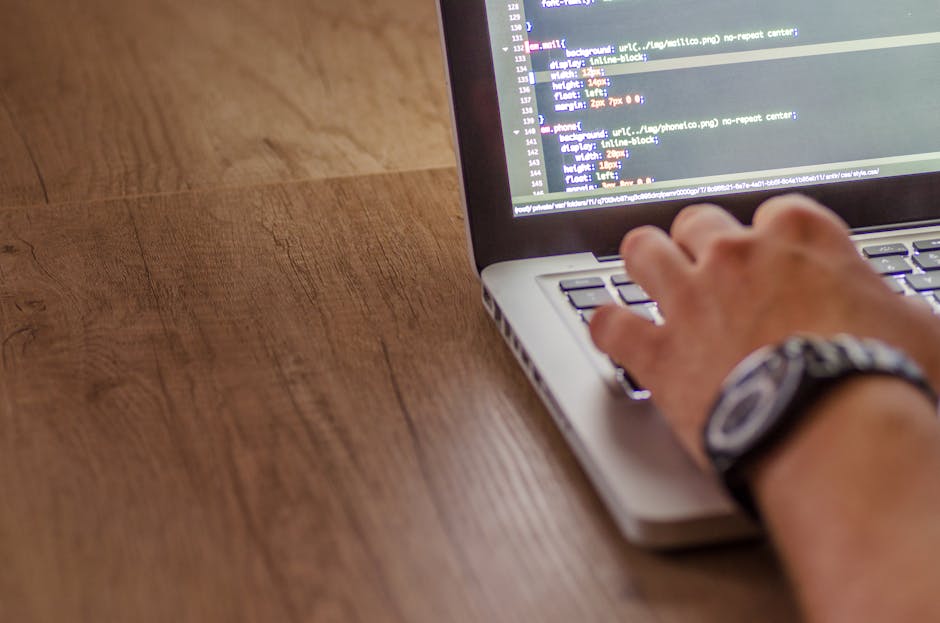 A person using a laptop placed on a wooden table, focusing on the screen which displays lines of code or a programming interface. The person's right hand is resting on the keyboard, with a wristwatch 