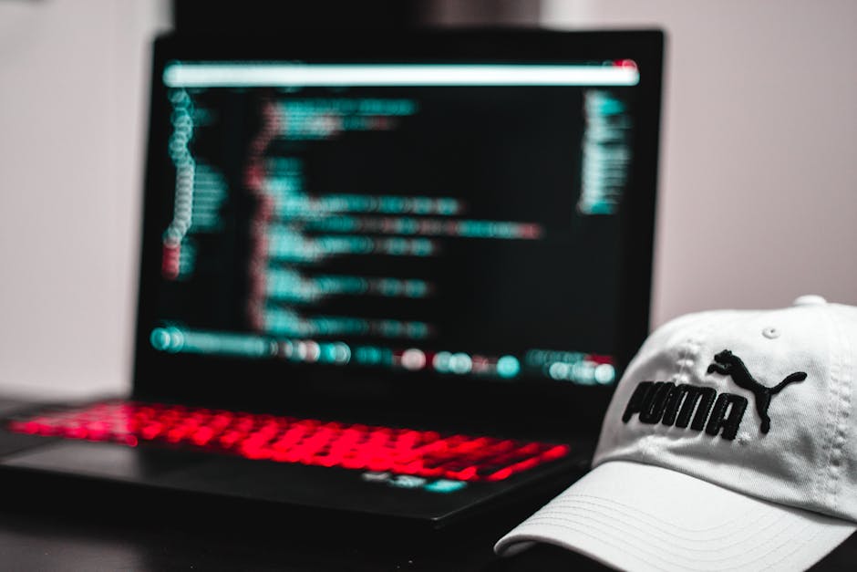 A person using a laptop placed on a wooden table, focusing on the screen which displays lines of code or a programming interface. The person's right hand is resting on the keyboard, with a wristwatch 